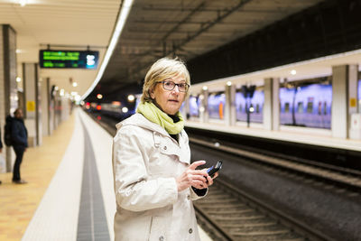 Senior woman looking away while standing with smart phone at subway station