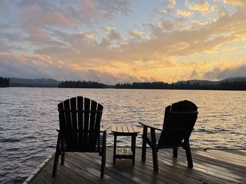 Empty chairs by lake against sky during sunset