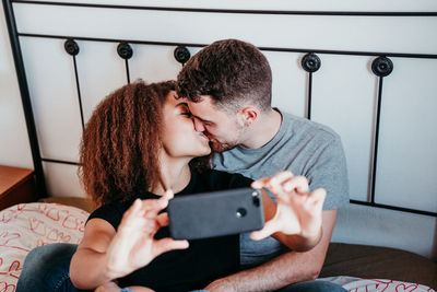 Laughing couple sitting on bed and kissing while taking selfie