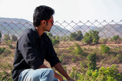 Man looking away while sitting on chainlink fence