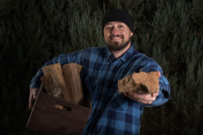 Portrait of smiling lumberjack holding wood and crate against trees at forest