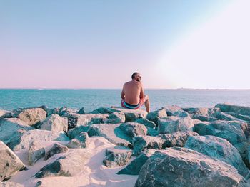 Rear view of shirtless man sitting on rocks by sea against sky