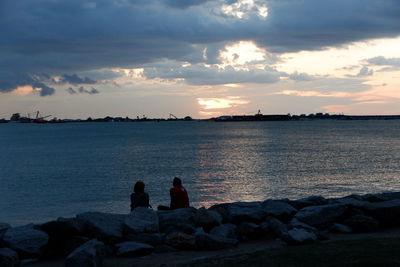 People sitting on rock by sea against sky during sunset