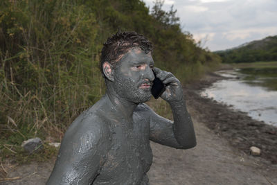 Man with mud on body against lake using mobile phone