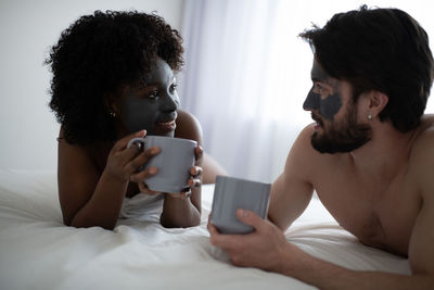 Multiracial couple with clay masks resting on bed