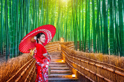 Mid adult woman holding red umbrella on steps amidst bamboo forest