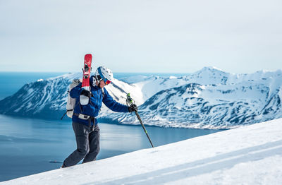 Man hiking uphill with skis and mountain and ocean behind him