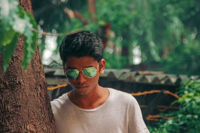 Portrait of young man wearing sunglasses while standing by tree trunk