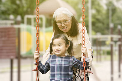 Senior woman playing with granddaughter sitting on swing in playground