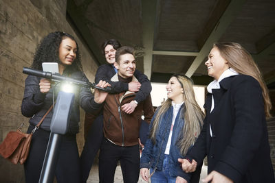 Smiling male and female teenage friends with electric push scooter on footpath below bridge