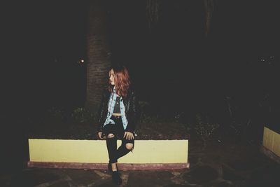 Full length of woman looking away while sitting on retaining wall at night