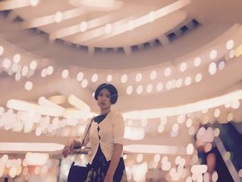 Low angle portrait of young woman against illuminated christmas lights at shopping mall