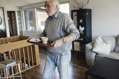 Senior man carrying serving tray while walking in living room at home