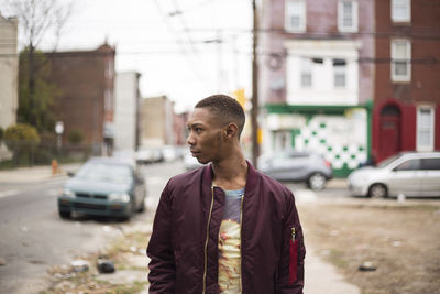 Young man wearing jacket standing on footpath against cars and buildings in city