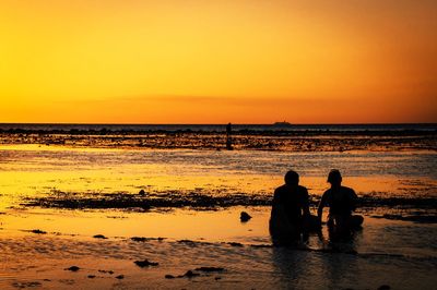 Silhouette men sitting on beach against sky during sunset