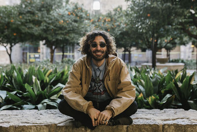 Smiling man with crossed legs sitting on retaining wall