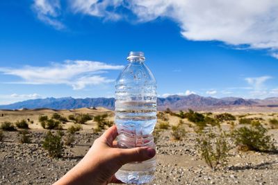 Cropped hand of person holding water bottle in desert against sky
