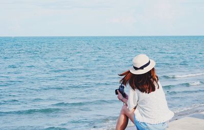 Rear view of woman with camera wearing hat while sitting at beach on sunny day