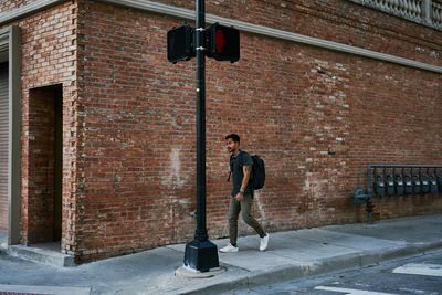 Hispanic male student in casual outfit with backpack walking along empty city street with bricked building on background