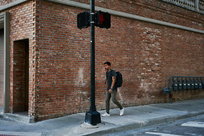 Hispanic male student in casual outfit with backpack walking along empty city street with bricked building on background
