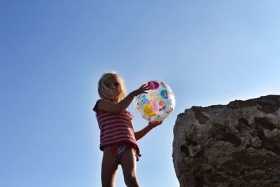 Low angle view of girl holding beach ball while standing by rock against clear sky