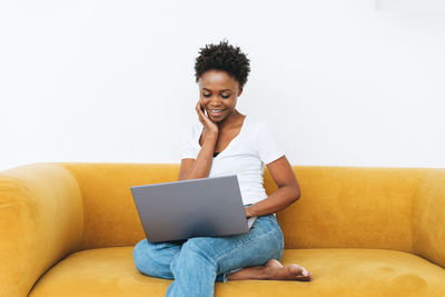 Beautiful african woman in white t-shirt and blue jeans using laptop on yellow sofa in bright