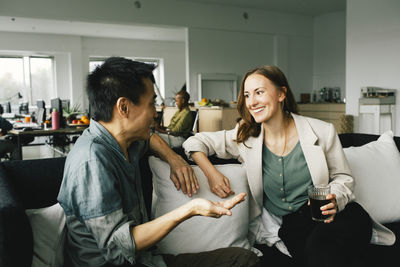 Happy female colleague holding drink while talking to businessman sitting on sofa at home