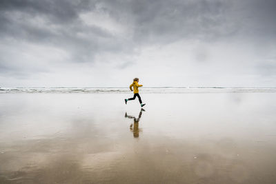 Side view of boy running at beach against cloudy sky