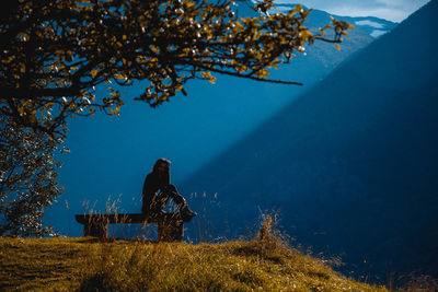 Woman sitting bench at field against mountain