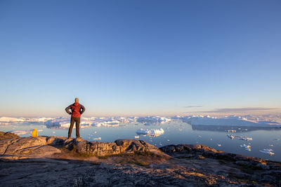 Man standing on rock by sea against clear blue sky