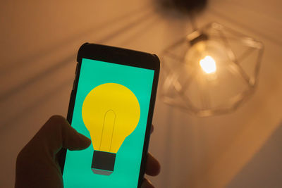 Cropped hand holding mobile phone against illuminated light bulb hanging on ceiling