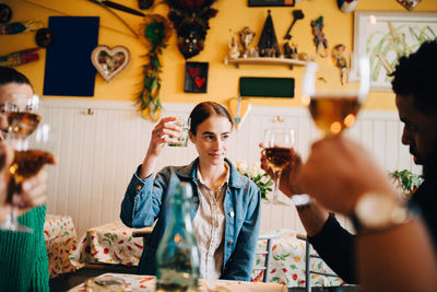 Young woman raising celebratory toast while sitting with multi-ethnic friends at dinner party in restaurant