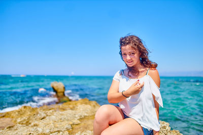 Portrait of young woman sitting on rock at beach against clear sky