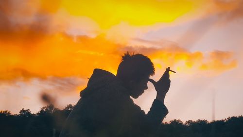 Silhouette man holding cigarette against sky during sunset