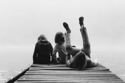 Rear view of friends with arm around sitting on pier by river during foggy weather