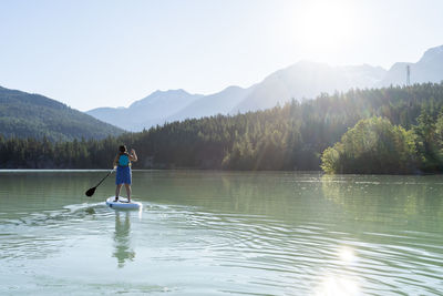 Barefoot woman in dress and life vest riding paddle board on calm lake water on sunny summer day in mountains in british columbia, canada