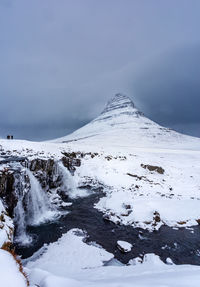 Scenic view of snowcapped mountain kirkjufell against stormy sky with waterfalls in the foreground 