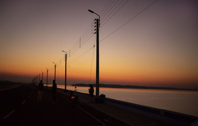 Silhouette electricity pylon by sea against sky during sunset