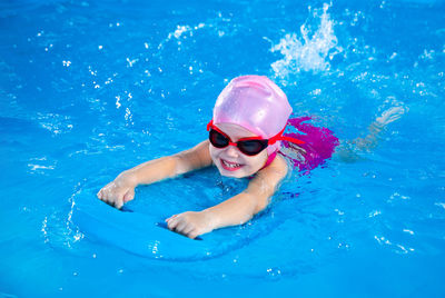 Smiling little girl learning to swim in indoor pool with flutterboard during swimming class