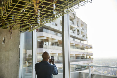 Mature man looking out of window while taking photo on smart phone