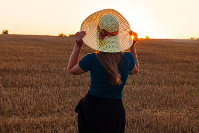 Rear view of woman wearing hat on field against sky during sunset in the wheat field countryside.