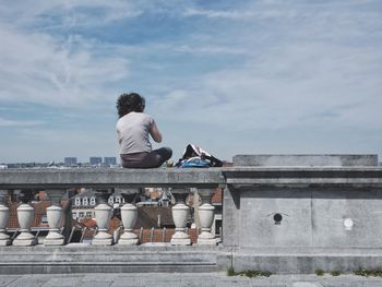 Rear view of woman sitting in railing against sky at city