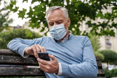 Senior man wearing mask using mobile phone while sitting on bench against tree