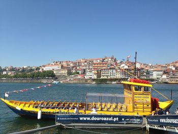 Boats moored in river by cityscape against clear blue sky