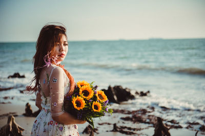 Woman with flowers standing on sea shore against sky