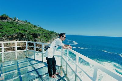 Man standing by railing against sea against clear blue sky