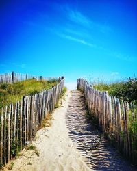 Wooden fence on footpath amidst field against blue sky