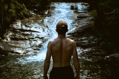 Rear view of shirtless man standing against waterfall