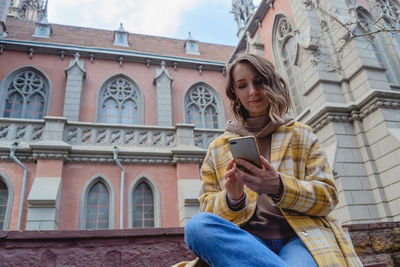 Happy millenial woman in a yellow coat in european city near historic building.