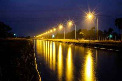 Illuminated street by river against sky at night